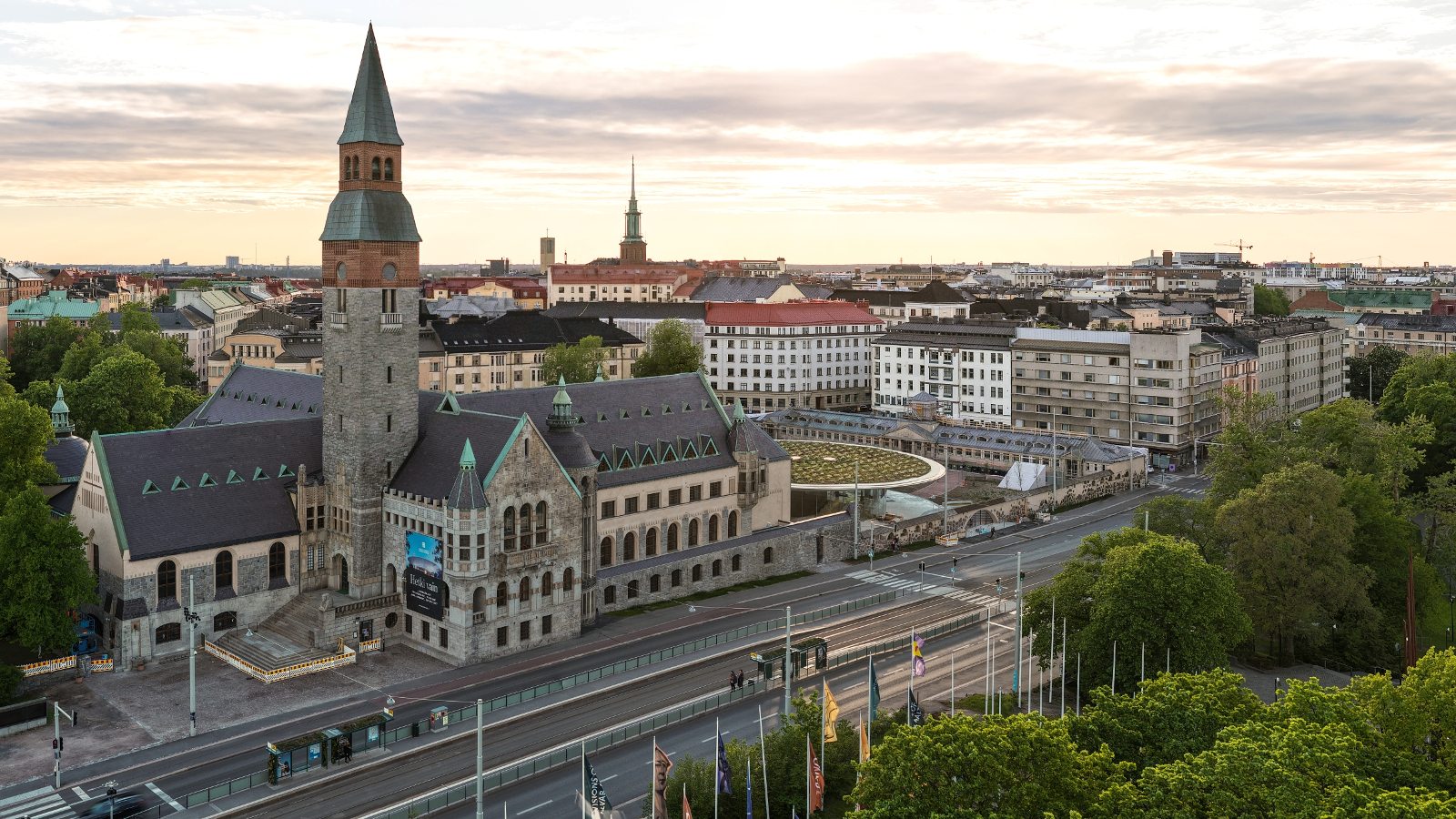 Kuvassa on Kansallismuseo ja sen ympäristö ilmasta käsin. Museo on harmaasta kivestä ja siinä on harmaa torni, jossa on puna-musta huippu. Taustalla on valkoisia taloja. Edessä on Mannerheimintie.