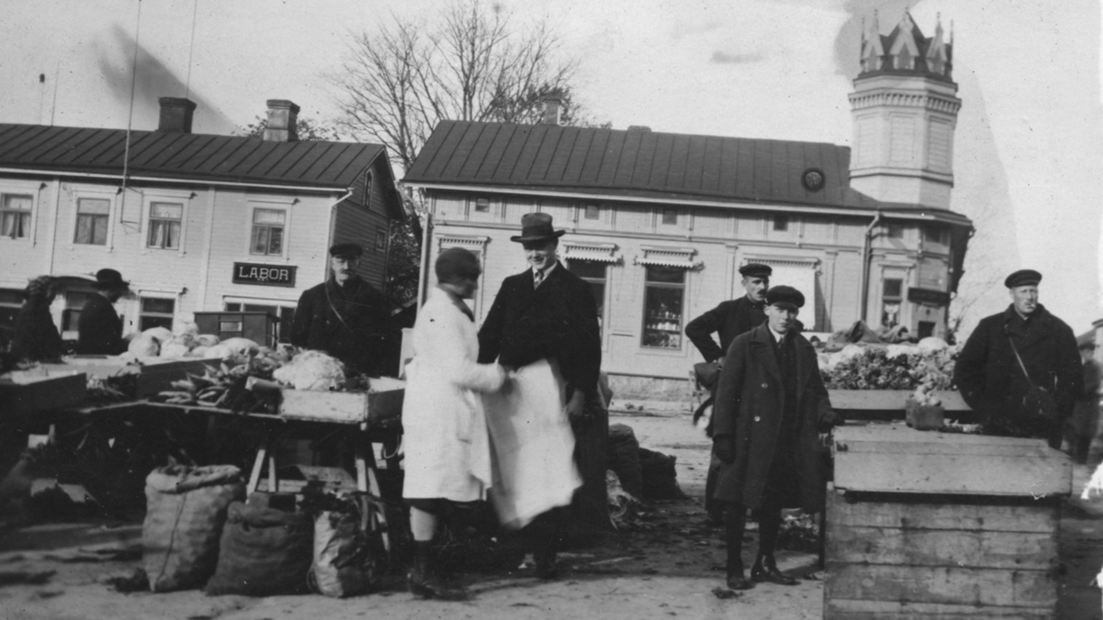 Kuvassa on etualalla torin myyntipöytä ja pöydän takana mies seisomassa. Pöydän vieressä seisoo nainen valkoisessa takissa ja mies mustassa hatussa ja lierihatussa. Oikealla on kaksi miestä ja välissä iso puulaatikko sekä sen vieressä mies. Taustalla on kaksi vaaleata, vanhaa puutaloa. Kuva on musta-valkoinen.