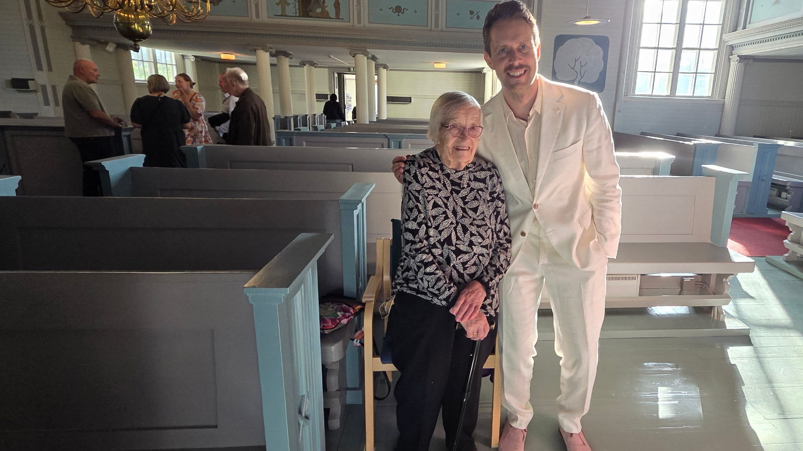 Kuvassa ovat seisomassa keskellä ja vasemmalla Marjatta Suur-Uski musta-valkopilkullisessa puserossa ja mustissa housuissa. Hänen oikealla puolellaan on Waltteri Torikka valkoisessa puvussa. Vasemmalla on turkooseja kirkon penkkäjä ja kauempana seisoo ihmisiä juttelemassa.