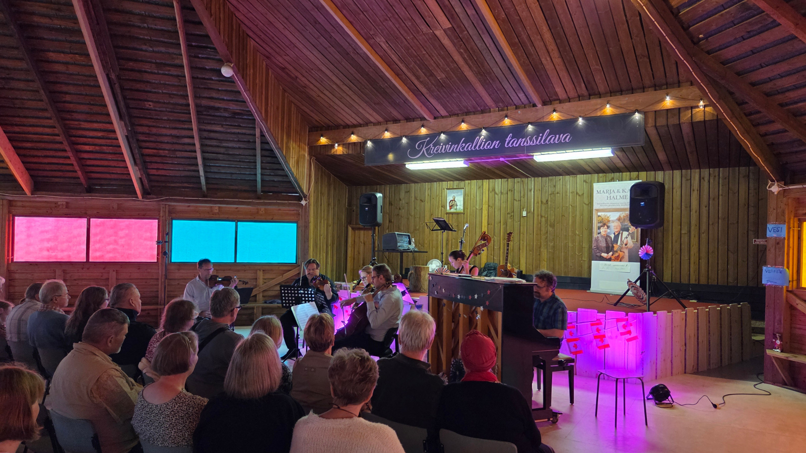 Kuvassa on vasemmalla yleisöä ja etualalla esiintymislavan alapuolella soittamassa Jukka Merjanen viulussa, Samuli Peltonen sellossa ja Maria Krykov kontrabassossa. Oikealla on piano ja Tuomas Turriago soittamassa. Taustalla näkyy pinkkiä valoa ja kattorakenteita , joissa on vinopalkkeja.