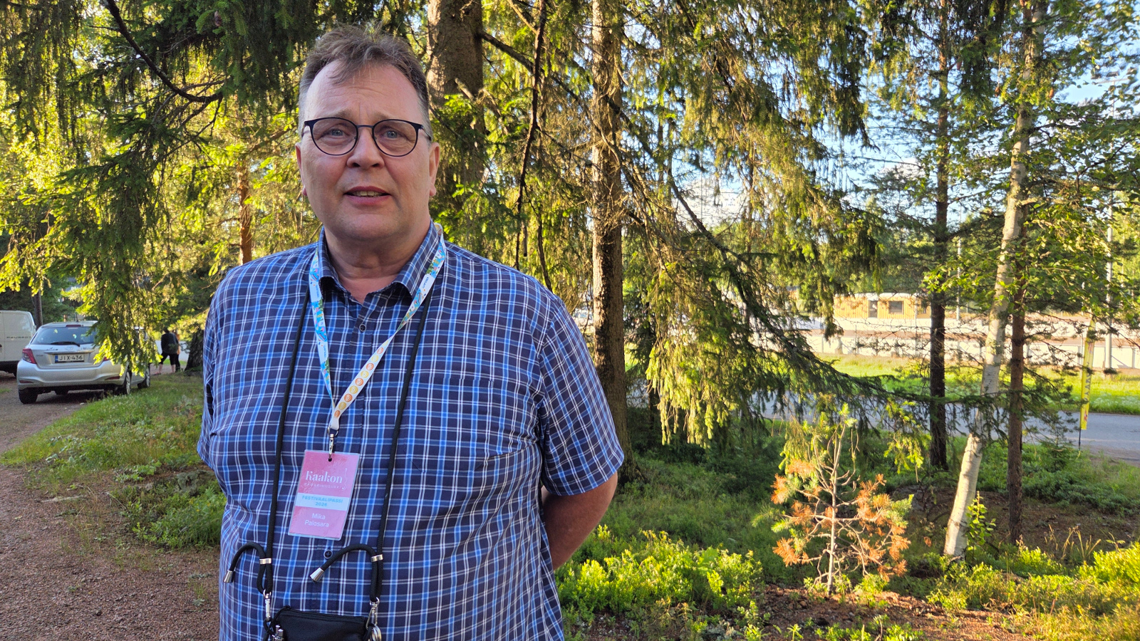 Kuvassa on Mika Palosara puolivartalokuvassa ja hänellä on sini-valkoruudullinen paita päällään. Taustalla on isoja kuusia oksineen.