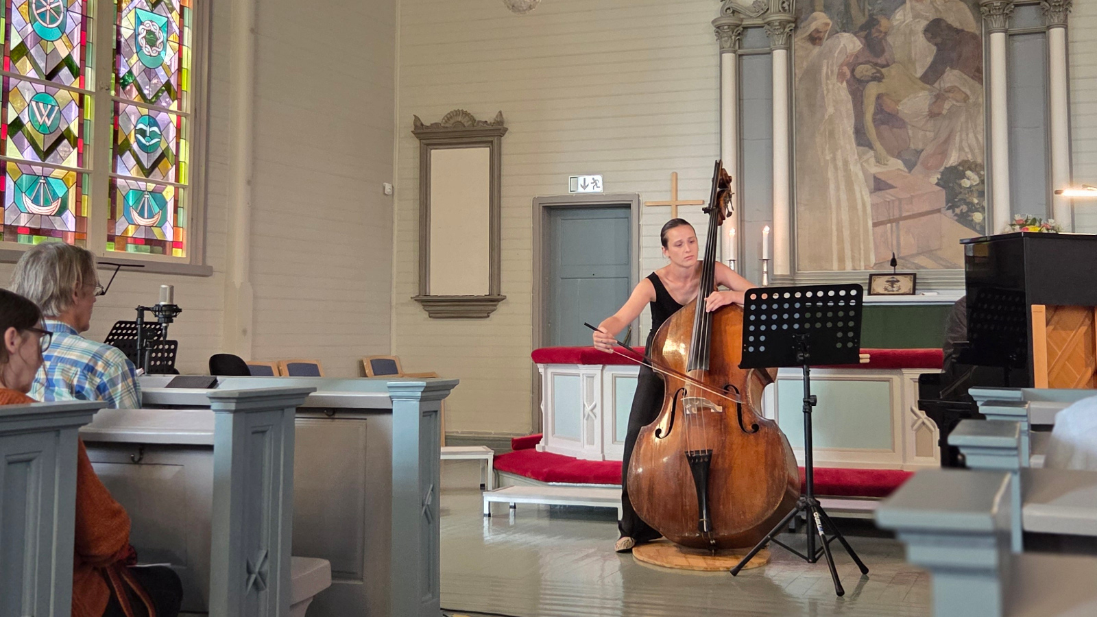 Kuvassa on keskellä Maria Krykov soittamassa kontrabasso ja oikealla näkyy pianoa. Vasemmalla on yleisöä turkooseissa penkeissä.