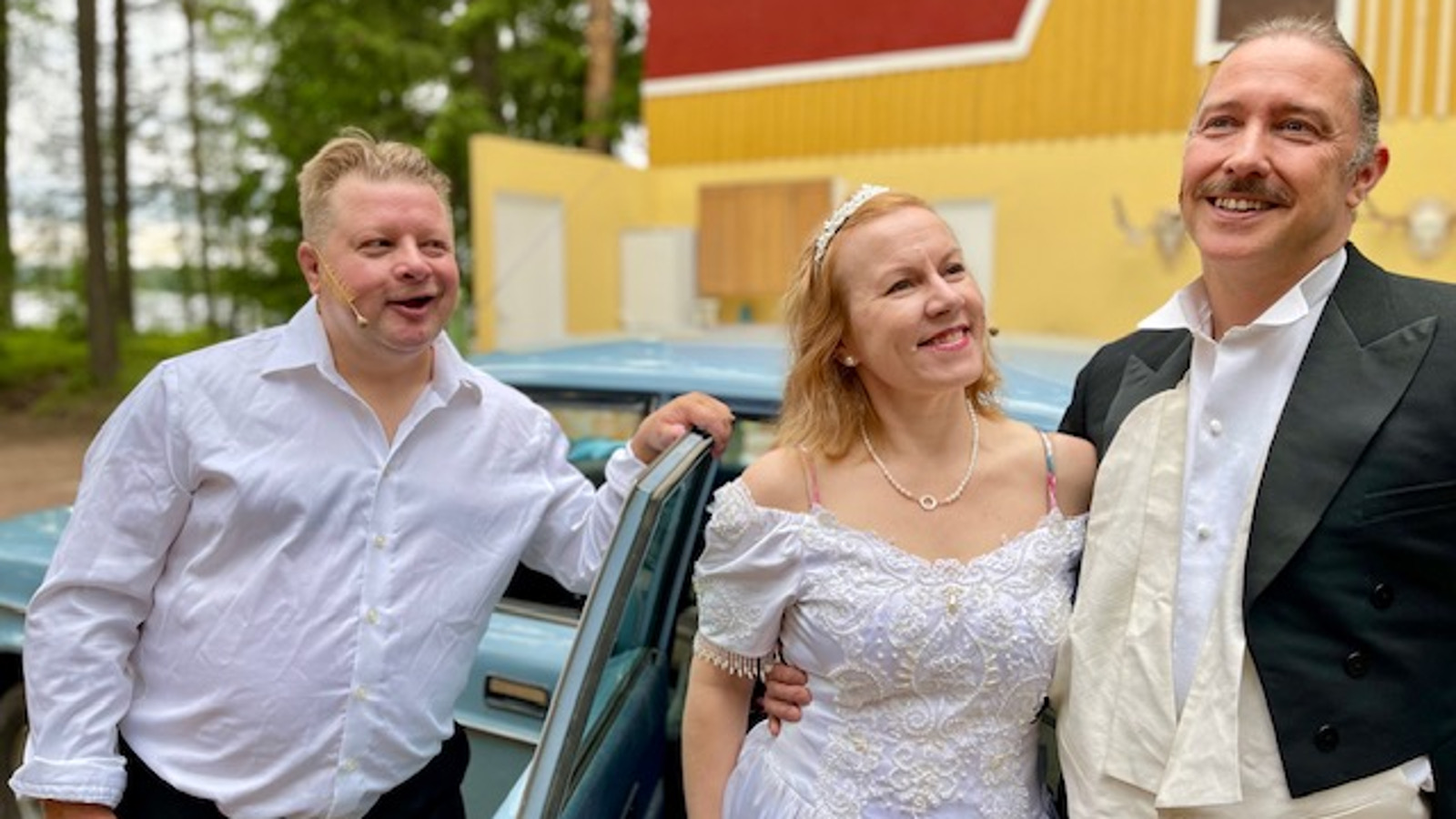 Kuvassa on vasemmalla mies valkoisessa paidassa ja oikealla pariskunta, jossa naisella on valkoinen morsiuspuku sekä miehellä tumma puku. Taustalla on vaalean sininen auto ja kelta-punainen rakennus.