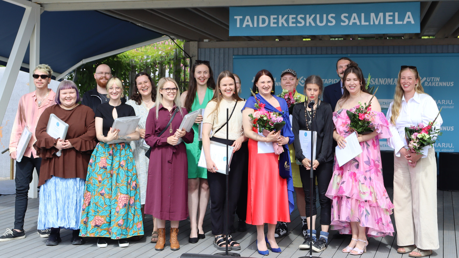 Kuvassa on seisomassa neljätoista taiteilija eri värisissä kesäisissä vaatteissa. Heidän yläpuolellaan on sininen kyltti, jossa lukee valkoisin kirjaimin Taidekeskus Salmela.   