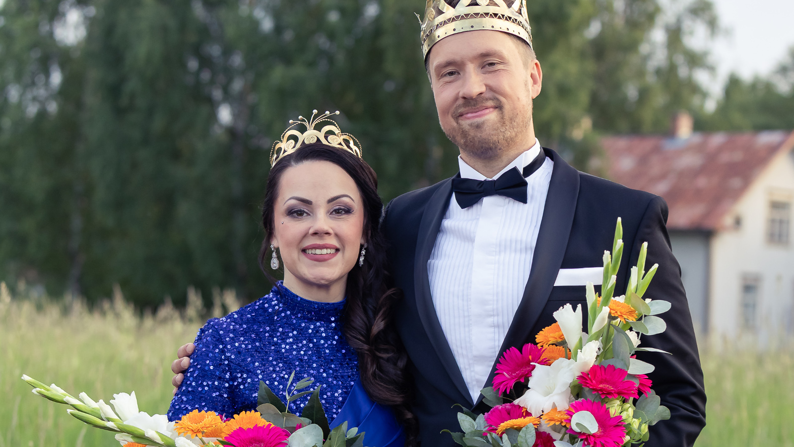 Kuvassa on vasemmalla Raija Mäntyniemi sinisessä mekossa ja vieressä oikealla Juho Punkeri mustassa takissa ja valkoisessa paidassa. Molemmilla on kukkakimput kädessään. Taustalla on metsää ja valkoinen talo.