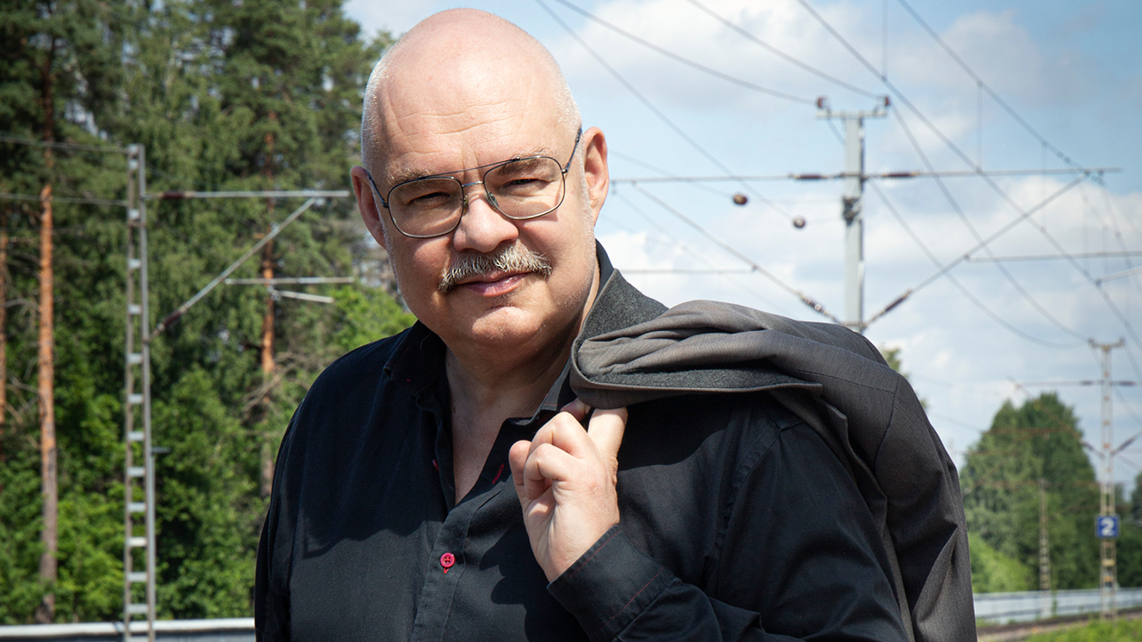Kuvassa on Mikko Roiha puolivartalokuvassa ja hänellä on musta paita ja musta takki vasemmalla olkapäällään. Taustalla näkyy metssä ja rautatien sähköjohtoja.