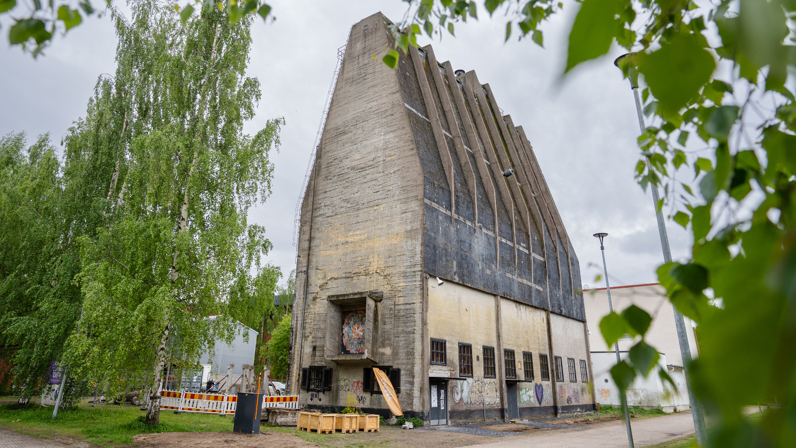 Kuvassa on keskellä harmaa, ylöspäin menevä harmaa siilo. Siinä on kattojuonteita tummalla katolla. Alhaalla on rivi ikkunoita. Molemmilla puolilla on vihreälehtisiä lehtipuita.