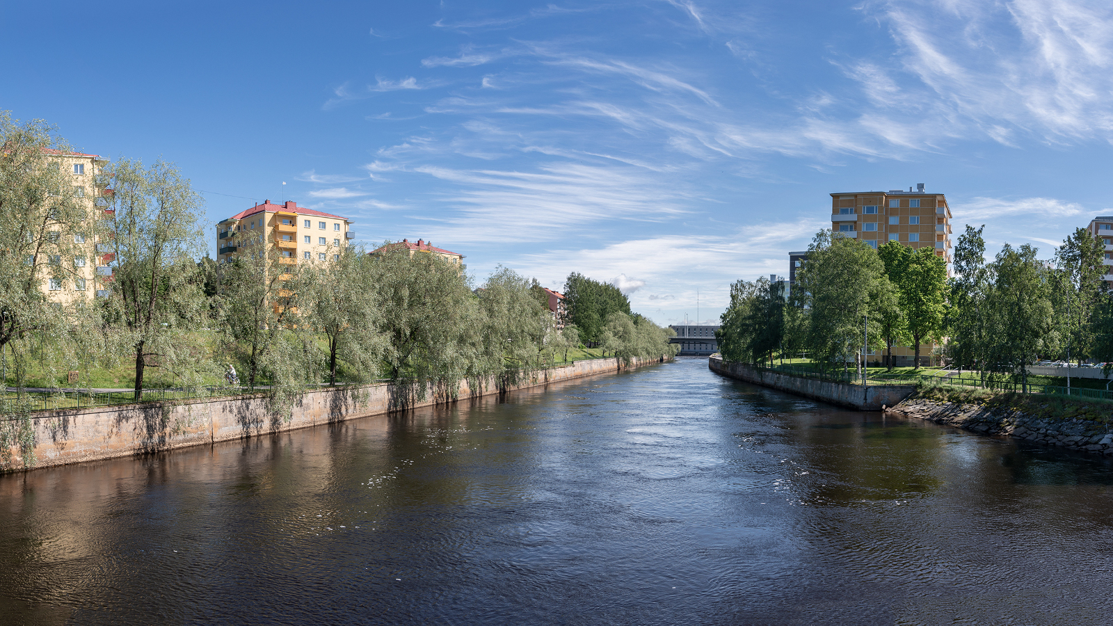 Kuvassa on keskellä joki ja sen molemmin puolin kerrostaloja, joiden edessä on lehtipuita. Takana on sinistä taivasta.