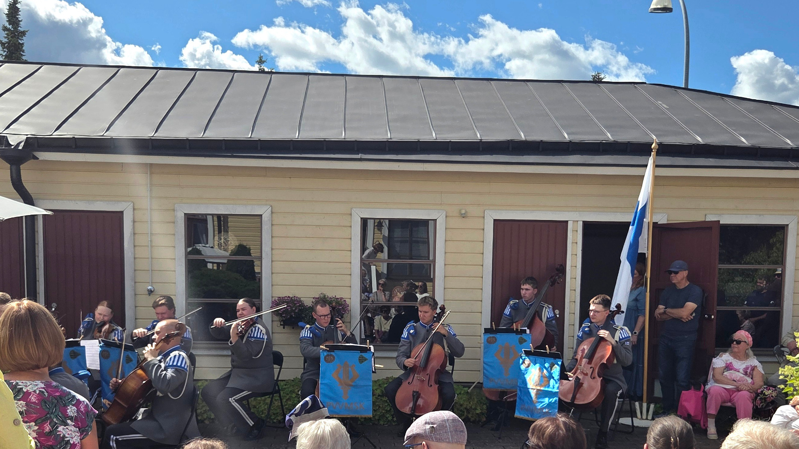 Kuvassa ovat harmaissa asuissa varusmiehet soittamassa selloilla ja viuluilla. Taustalla on keltainen rakennus, jossa on isoja ikkunoita. Oikealla on Suomen lippu.