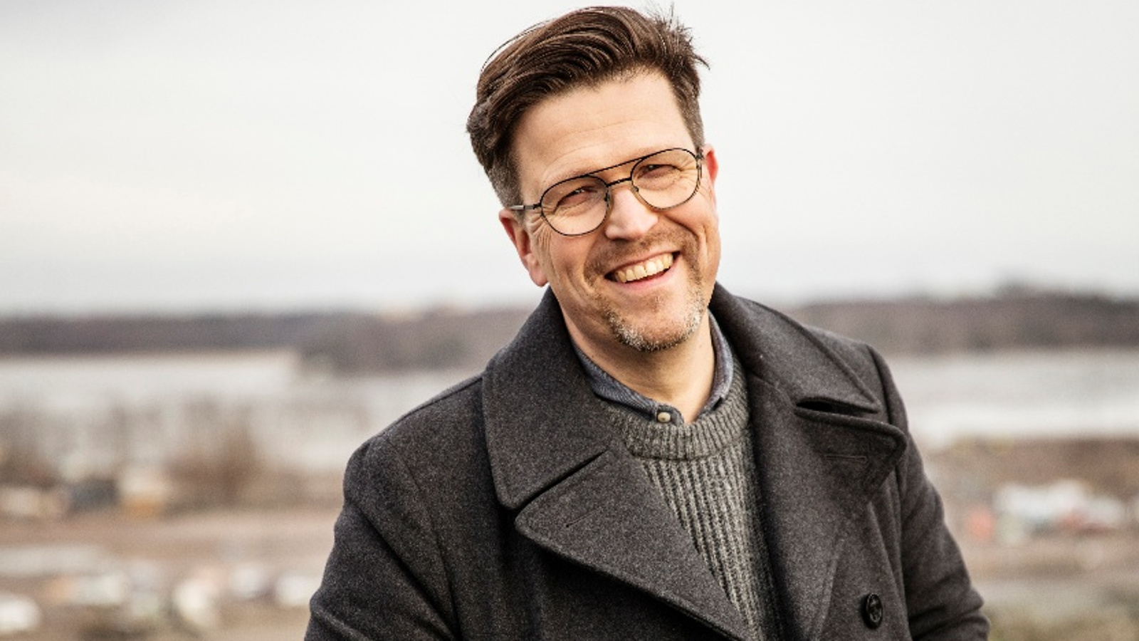 Kuvassa on Klaus Härö puolivartalokuvassa ja hänellä on ruskea takki ja sen alta näkyy harmaan ruskeata villapaitaa. Taustalla on epätarkkaa syysmaisemaa.
