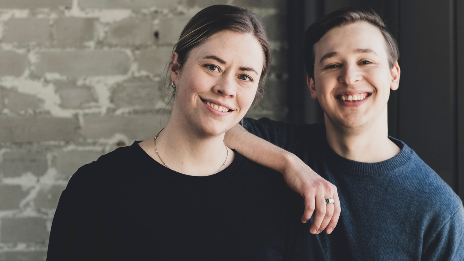 Kuvassa on vasemmalla Lotta Fors ja oikealla Jakob Norrgård. Hän pitää oikeata kättään Lotta Forsin olkapäällä. Heillä molemmilla on mustat t-paidat päällään. Taustalla on harmaan valkeaa tiiliseinää.