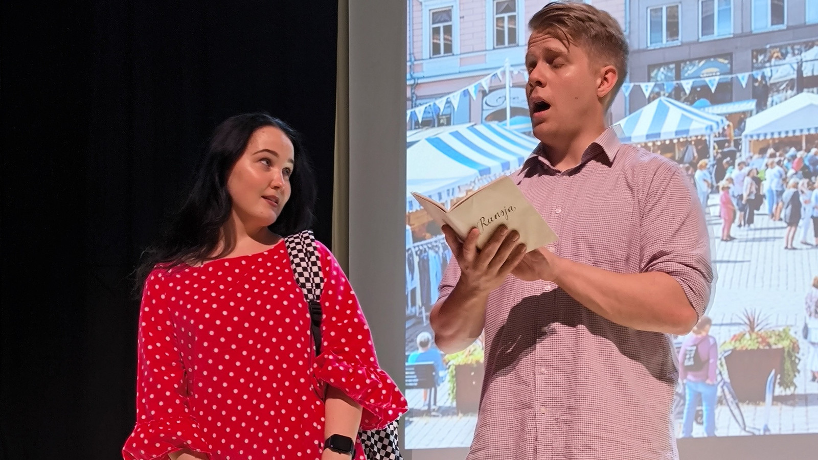 Kuvassa on vasemmalla nuori nainen punaisessa mekossa. Oikealla on nuori mies vaalean punaisessa paidassa ja pitää kirjaa kädessään ja laulaa.