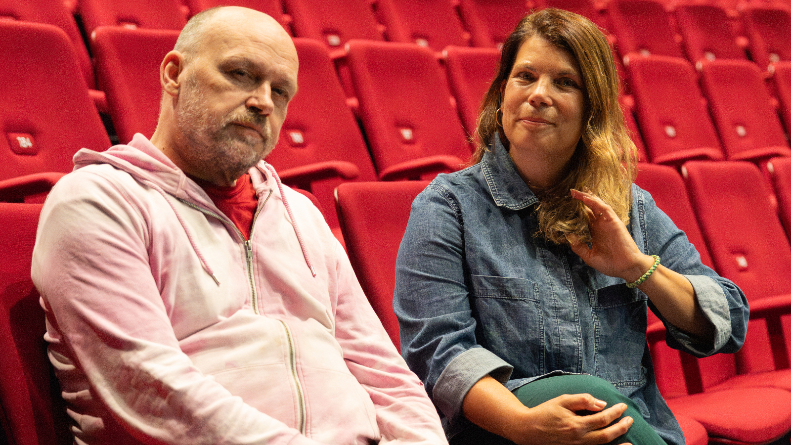 Kuvassa ovat vasemmalla Daniel Wetzel vaalean punaisessa paidassa ja Helgard Haug sinisessä puserossa. He istuvat punaisilla istuimilla ylenevässä teatterin katsomossa.