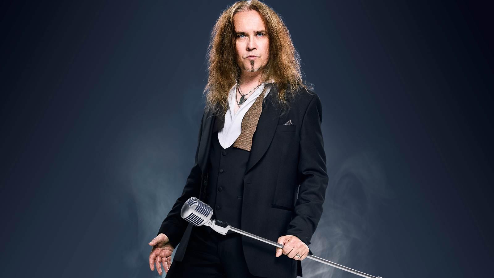 Kuvassa on keskellä on Jarkko Ahola seisomassa ja hänellä musta takki sekä valkoiset kaulukset. Hänellä on jalallinen mikrofoni oikeassa kädessään. Tausta on tumma.