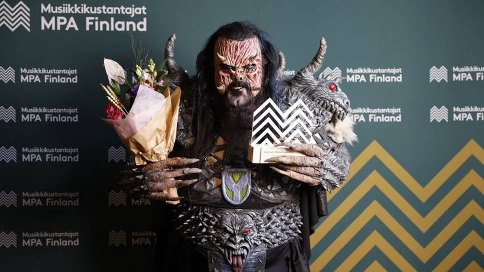 Kuvassa on Lordi monsteriasussaan, jossa on juovikas naamari ja olkapäistä lähtevät sarvet. Pukuun kuuluvat musta, paksu vyö, jossa on hirviön naama. Käsissään Lordilla on kukat ja palkinto.