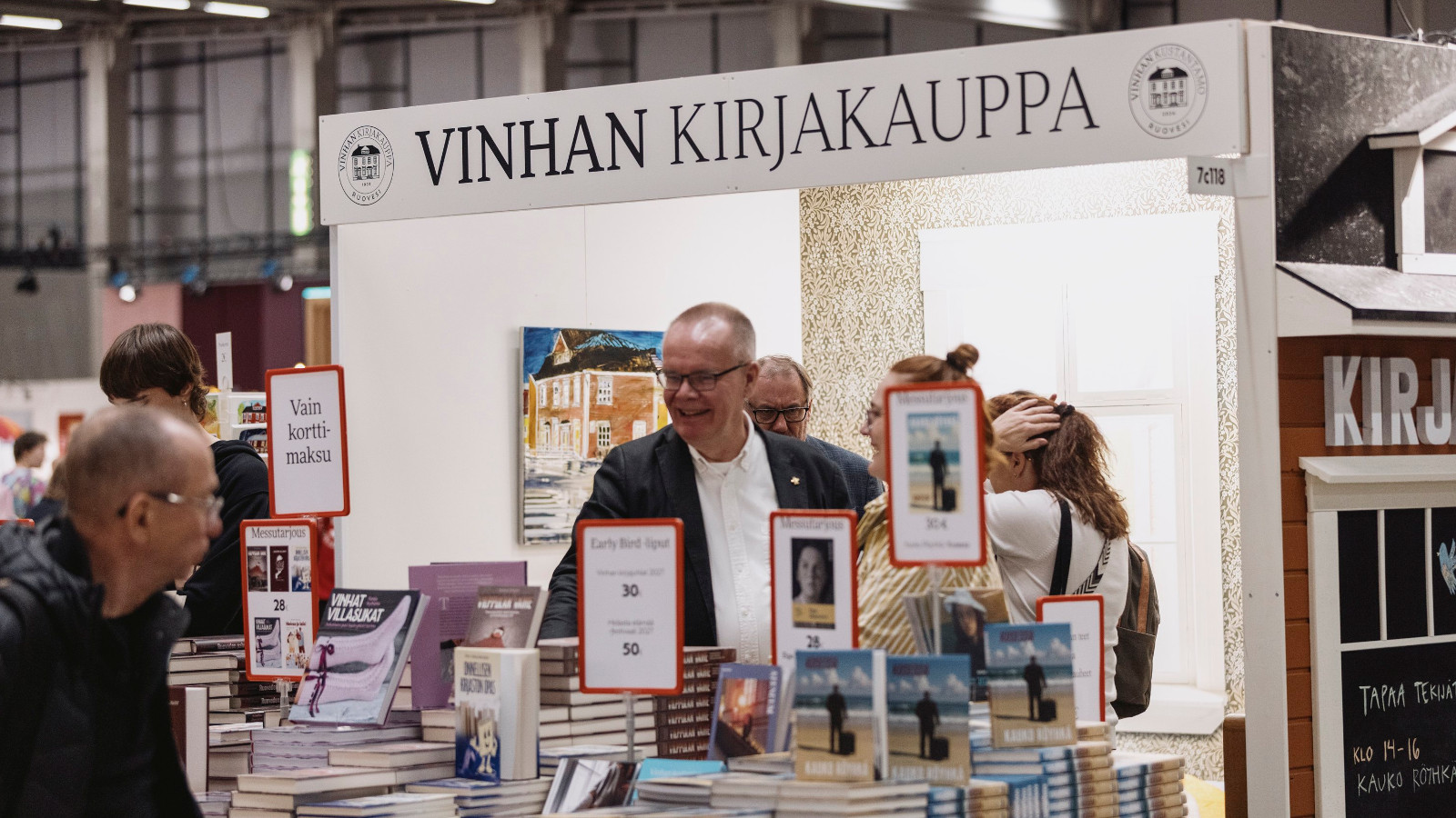 Kuvassa on Vinhan kirjakaupan osasto ja yläosassa on kyltti. Etualalla on kirjoja ja takana kaksi henkilöä ja vasemmalla osastolla kävijä.