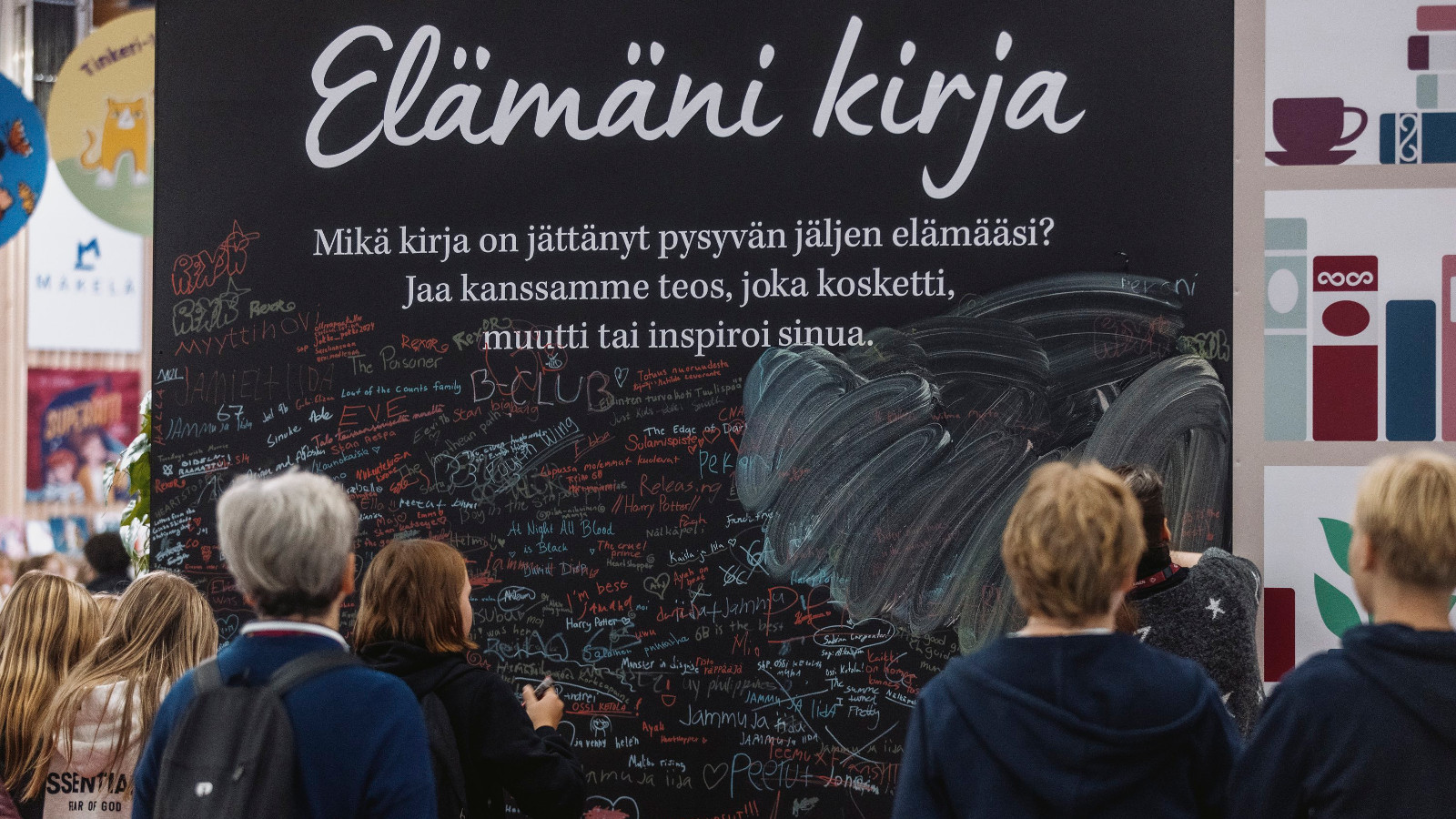 Kuvassa on seinämä, jossa lukee kirjoituskirjaimin Elämäni kirja ja sen alla pientä tekstiä mustalla pohjalla. Edessä on ihmisiä katselemassa seinää.