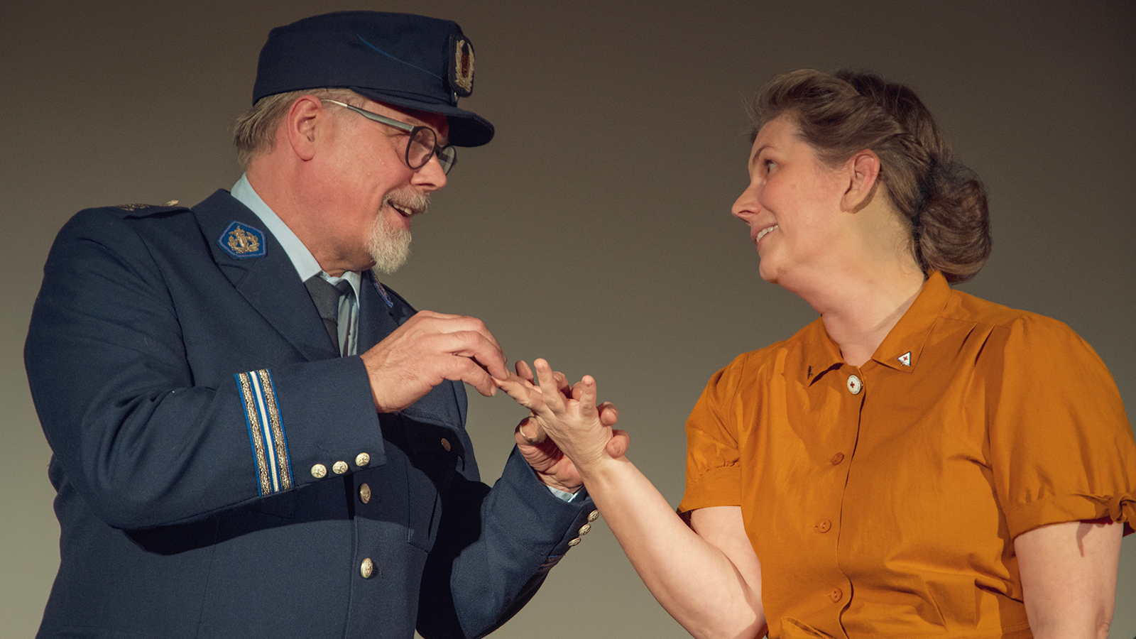Kuvassa on vasemmalla puolivartalokuvassa mies poliisipuvussa ja hän pitää kädestä naista, jolla on tumman oranssi pusero.