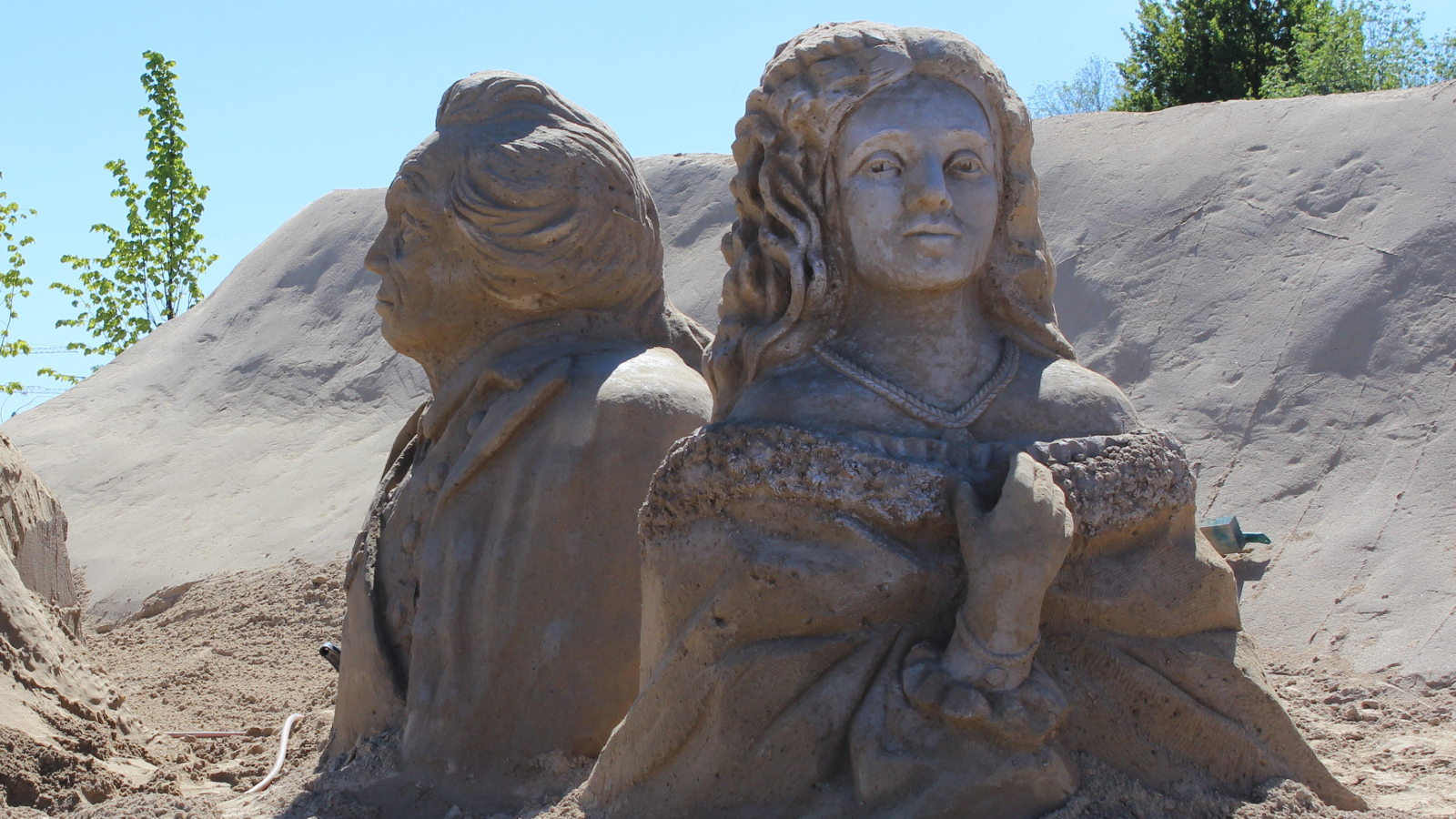 Kuvassa keskellä on kaksi suurehkoa hiekkaveistosta, joissa aatelisasuissa oleva nainen ja mies rintakuvina.  Nainen katsoo suoraan kuvan suuntaan ja mies vasemmalla poispäin.  Veistokset ovat hiekan päällä ja takana vasemmalla näkyy sinistä taivasta.