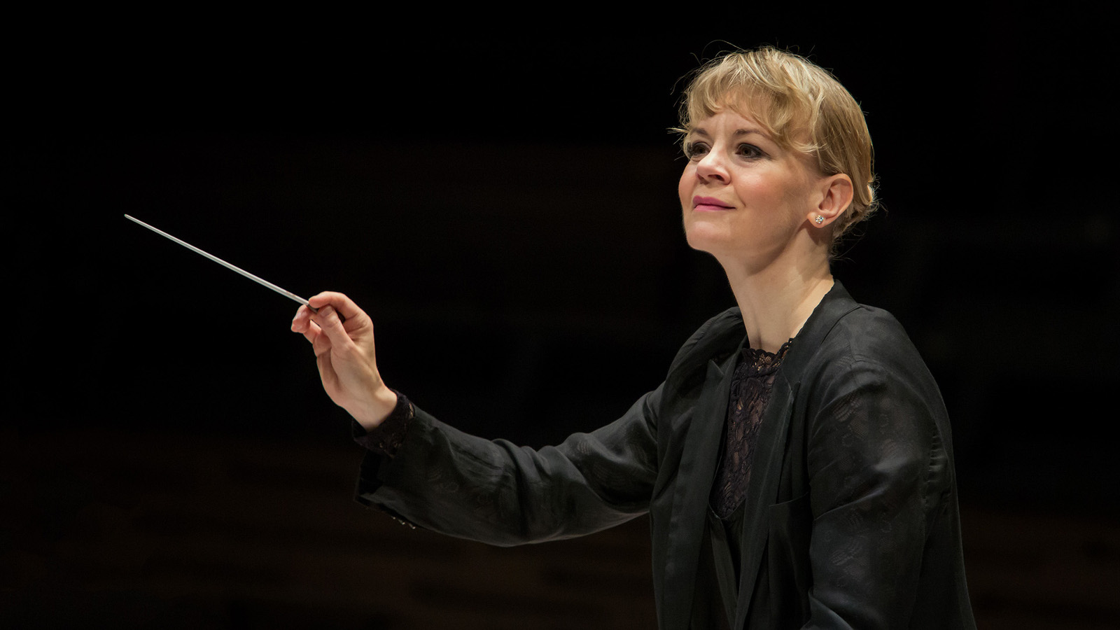 Kuvassa Susanna Mälkki johtamassa orkesteria mustassa puserossa ja kädessään tahtipuikko. Tausta on musta ja kuvassa näkyy vain Susanna Mälkki. 