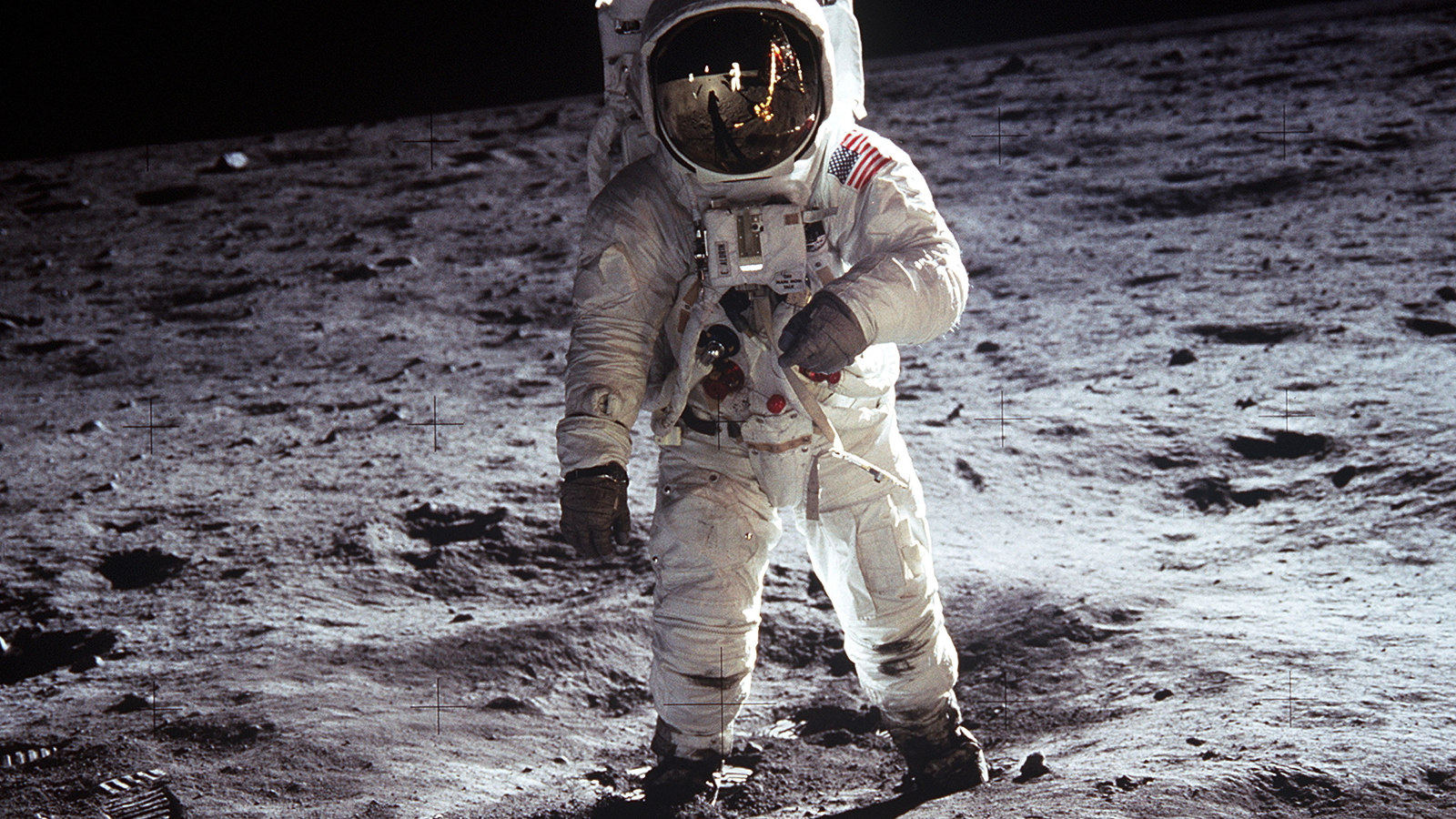 Kuvassa on astronautti Buzz Aldrin kävelyllä valkoisessa avaruuspuvussa, jossa on USA:n lippu hihassa.  Kuun pinta näkyy tummana ja vaaleana kivisenä pintana.