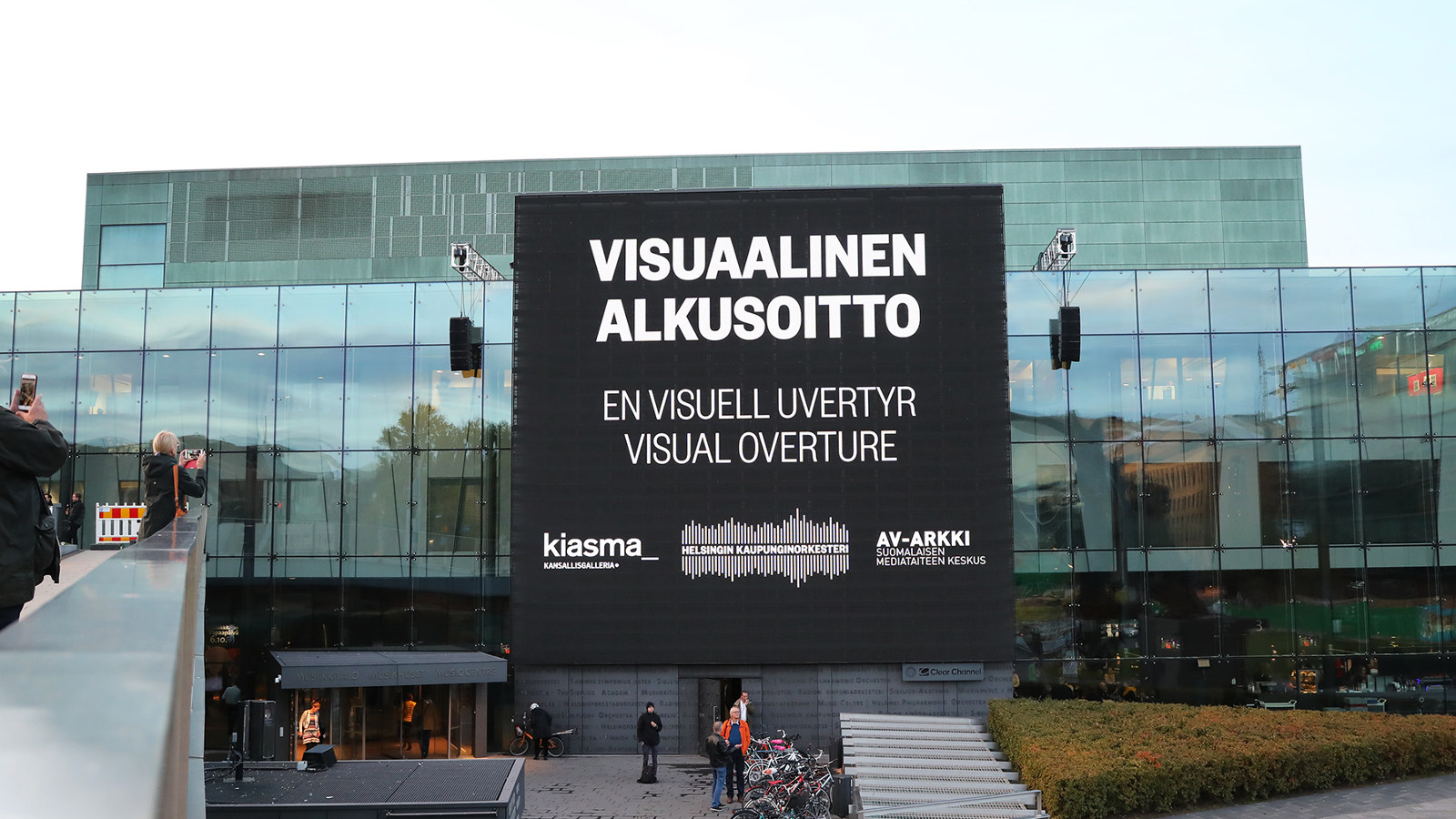 Kuva on Musiikkitalosta harmaana päivänä.  Musiikkitalon isolla näytöllä lukee Visuaalinen alkusoitto, en visuell uvertyre,visual overture. Alla on yhteistyökumppaneiden Kiasma, Helsingin kaupunginorkesterin ja Av-arkin logot.