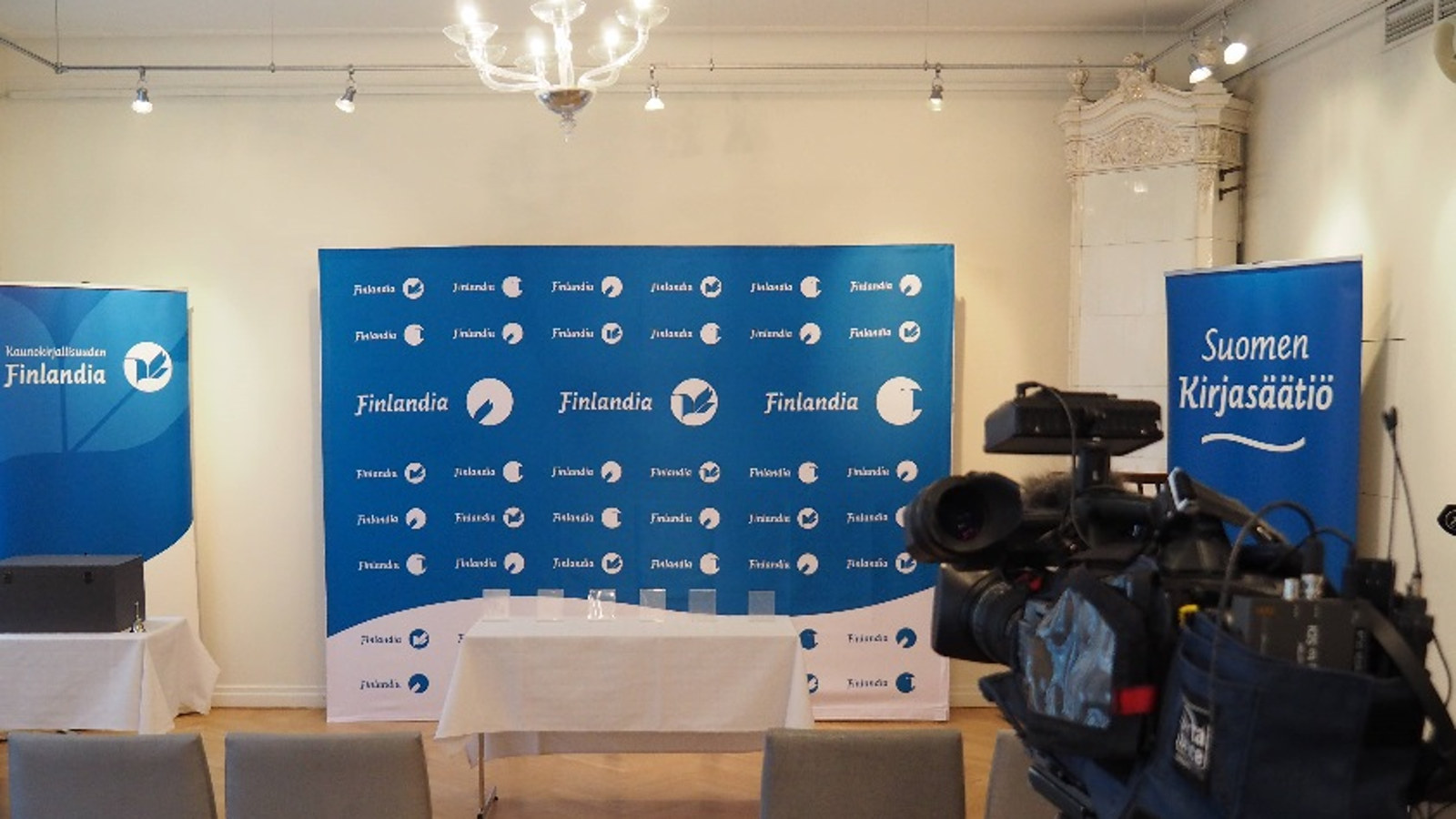 Kuvassa on Finlandia-palkintosasettelu.  Kuva on huoneesta, jossa on taustalla sini-valkoinen Finlandia-palkintojen logojen täyttämä taulu ja Suomen Kirjasäätiön Finlandia-palkinnon roll-upit.  Etualalla on tuoleja ja  teknistä välineistöä.