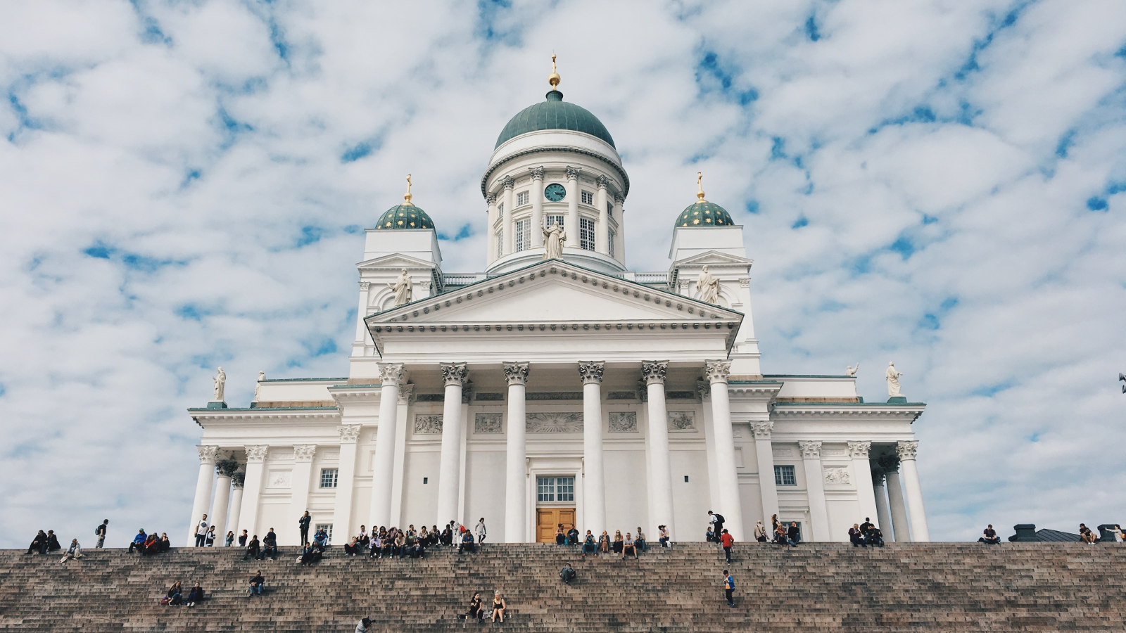 Kuvassa on Helsingin tuomiokirkko, joka on väriltään valkoinen.  Taustalla on sinistä taivasta ja siinä on valkoisia pieniä pilviä.  Alempana on ylempiä portaita ja niillä istuu ihmisiä siellä täällä.