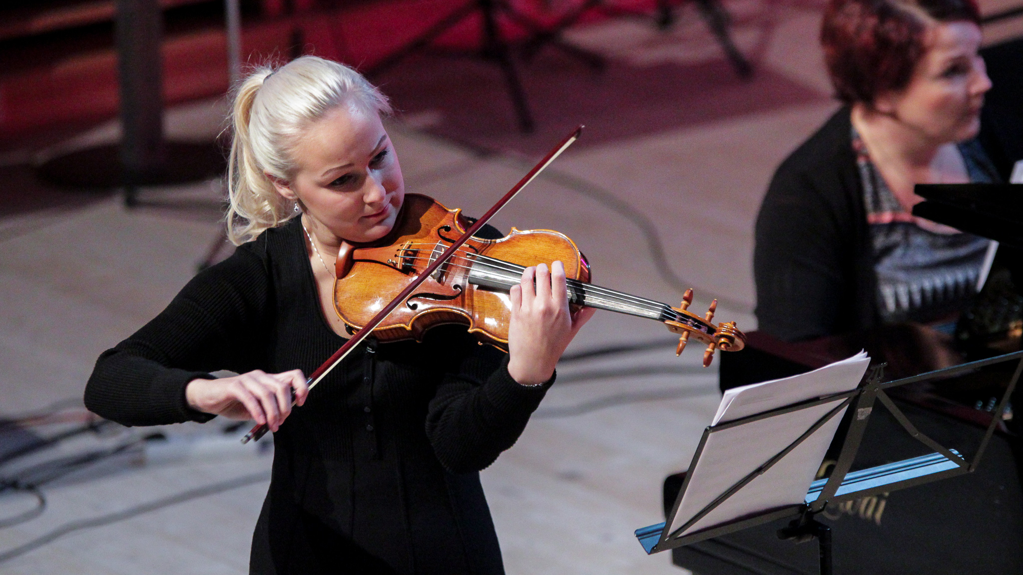 Kuvassa on nuori vaaleatukkainen nainen soittamassa viulua.  Kuvassa näkyy myös toinen soittaja istumassa.  