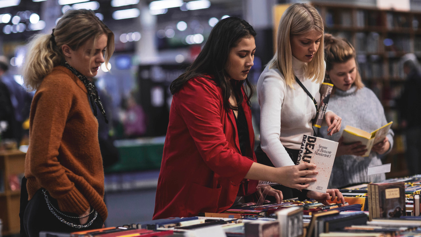 Kuvassa on kolme nuorta naista katsomassa kirjoja Helsingin Kirjamessuilla. Heidän edessään on täysi kirjapöytä.