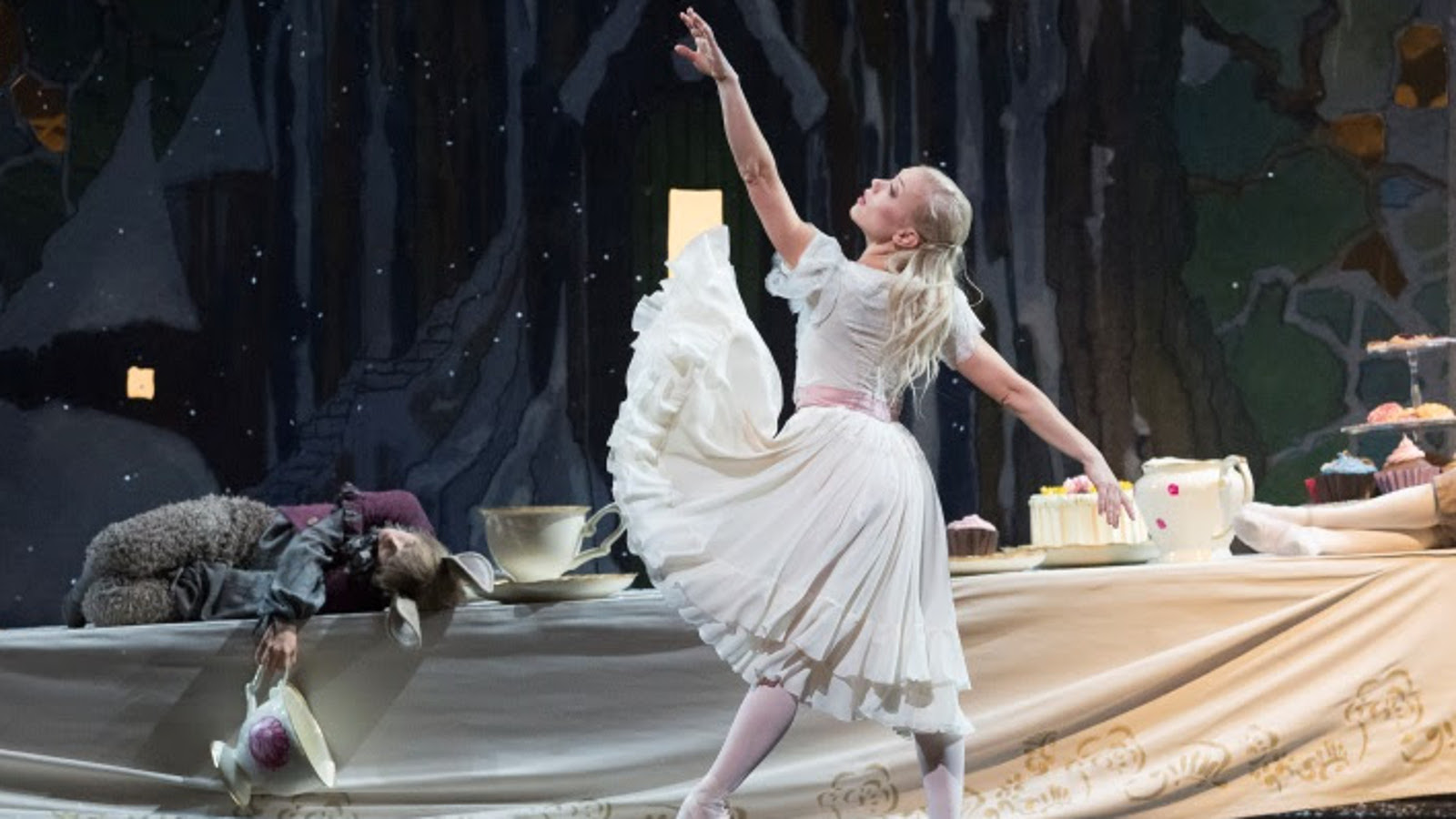 Kuvassa on Liisa Ihmemaassa baletin Liisa, Linda Haakana.  Hänellä on vaalea mekko ja hän osoittaa vasemmalla kädellään ylöspäin.  Lattialla makaa kani vasemmalla harmaassa puvussa.  Taustalla on tumman harmaa verho ja oikealla lattialla on kakkuja.