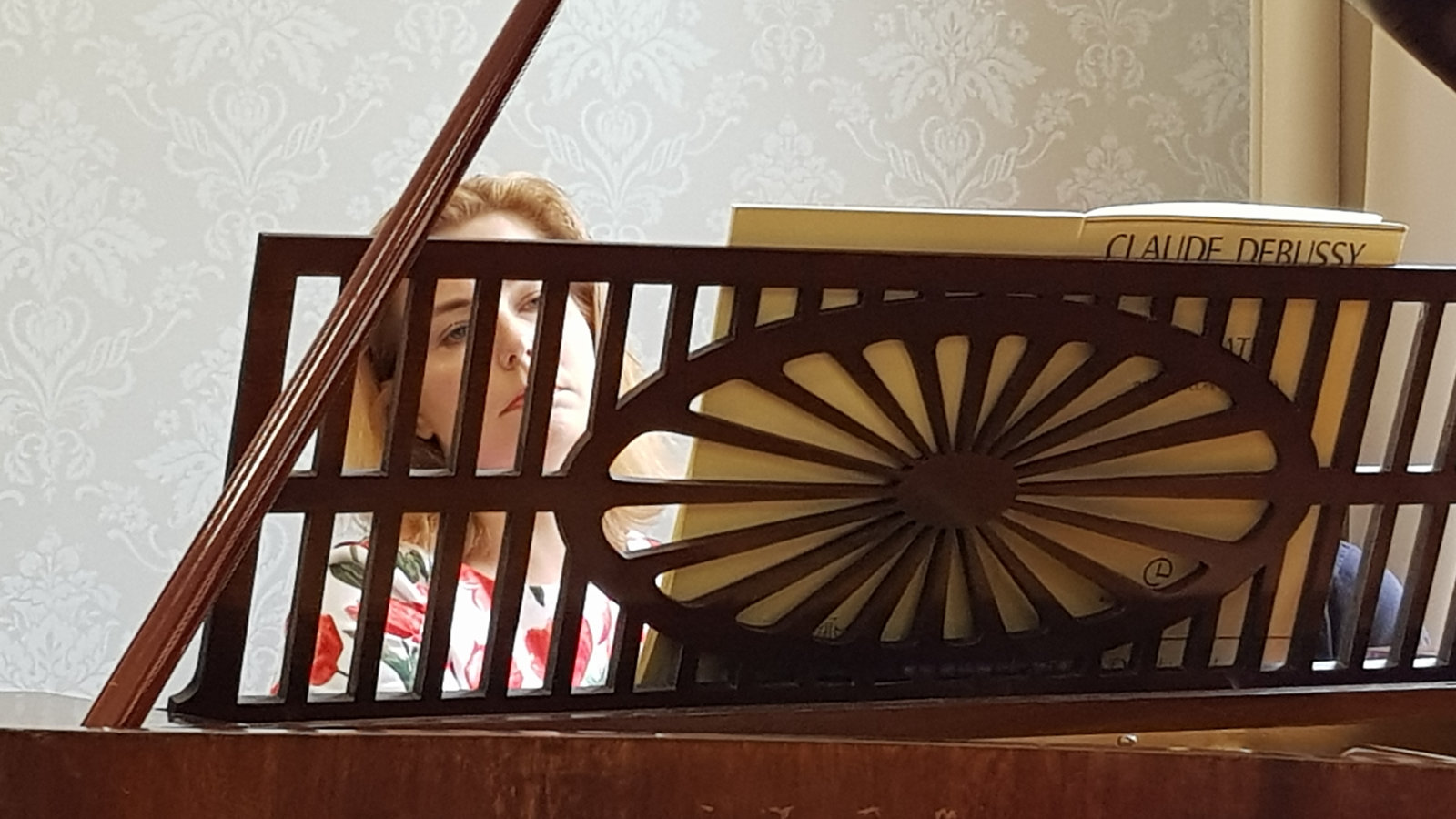 Kuvassa on Liisa Malmivaara soittamassa pianoa ja hänen kasvonsa näkyvät ruskearuudullisen pianon nuottitelineen takaa.  Piano on vanha.  Malmivaaralla on vaaleat hiukset.