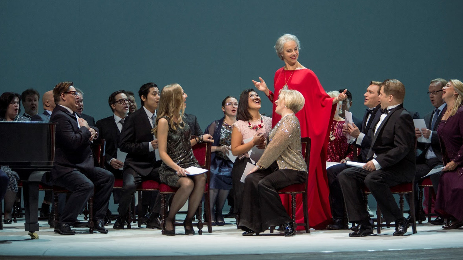 Kuvassa on mustissa puvuissa olevia ihmisiä, jotka istuvat.  Heidän keskellään seisoo Anne Sofie von Otter punaisessa, pitkässä puvussa.