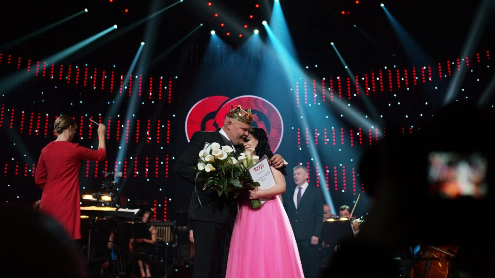 Kuvassa ovat tangokuninkaalliset 2019, vasemmalla Johannes Vatjus tummassa puvussa ja oikealla Pirita Niemenmaa vaaleanpunaisessa puvussa.  He halaavat toisiaan lavalla ja takana on punamusta tausta sekä sinisiä  valojuovia.  Vasemmalla on selin punaisessa mekossa oleva orkesterinjohtaja.