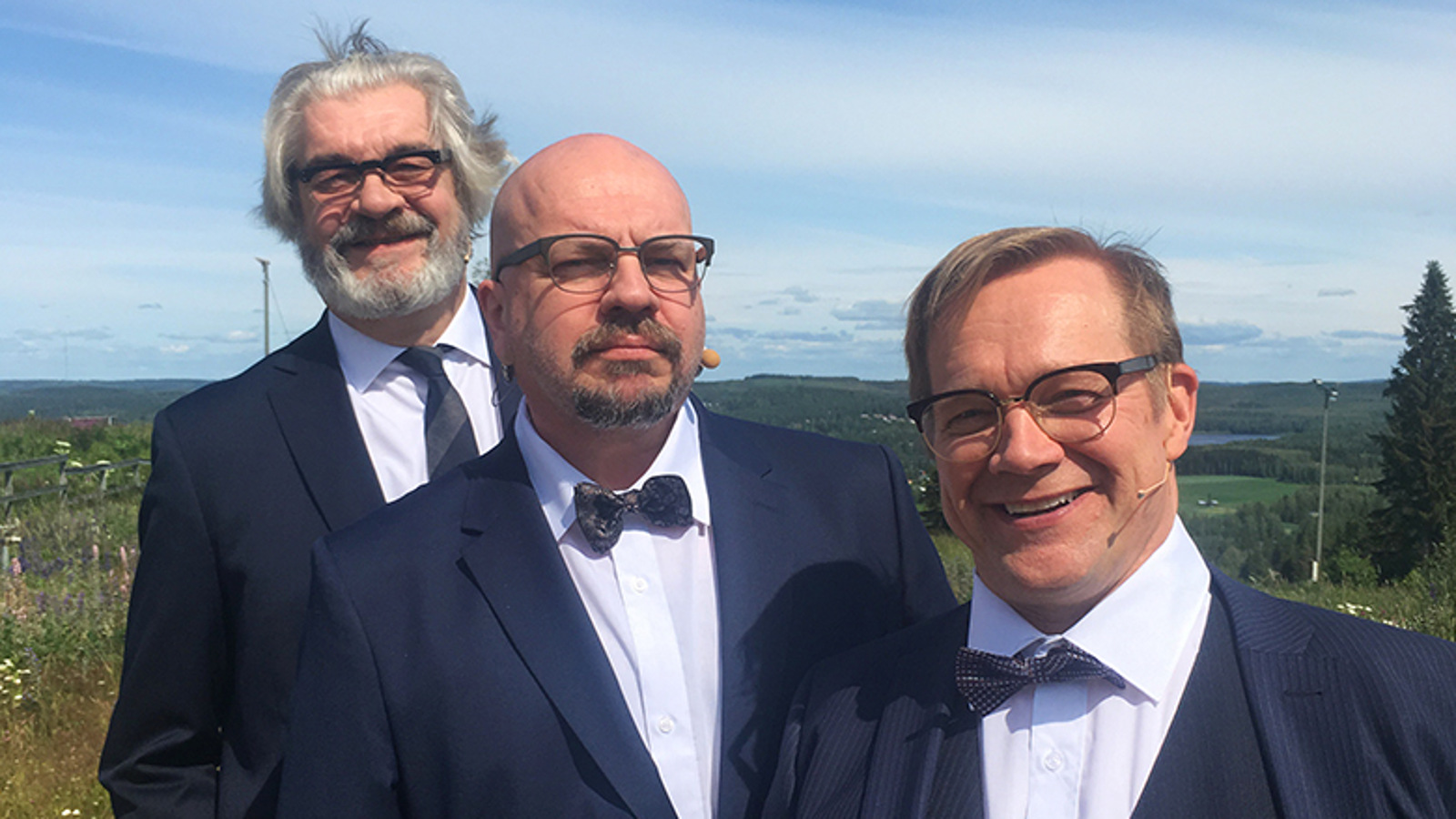 Kuvassa ovat Jussi Lampi, Timo Rautiainen ja Puntti Valtonen puolivartalokuvassa ja limittäin rinteessä.  Heillä on tummat puvuntakit päällä ja valkoiset paidat sekä tummat solmiot.  Keskellä olevalla Timo Rautiaisella on rusetti.  Taustalla on sininen, kirkas taivas.