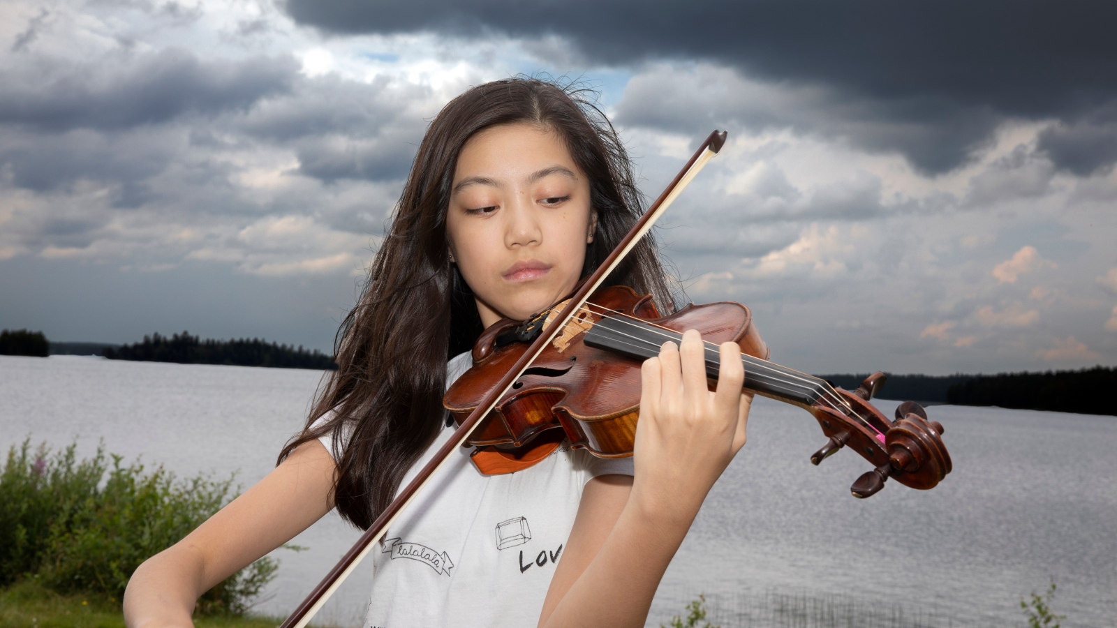 Kuvassa on nuori viulistioppilas Hannah Tam, jolla on mustat pitkät hiukset ja  soittaa viulua. Hänestä on puolivartalokuva ja  päällään valkoinen t-paita, jossa lukee mm. love. Taustalla on järvimaisemä ja ylimpänä tumman harmaa pilvi ja muu taivas oli puolipilvinen.   