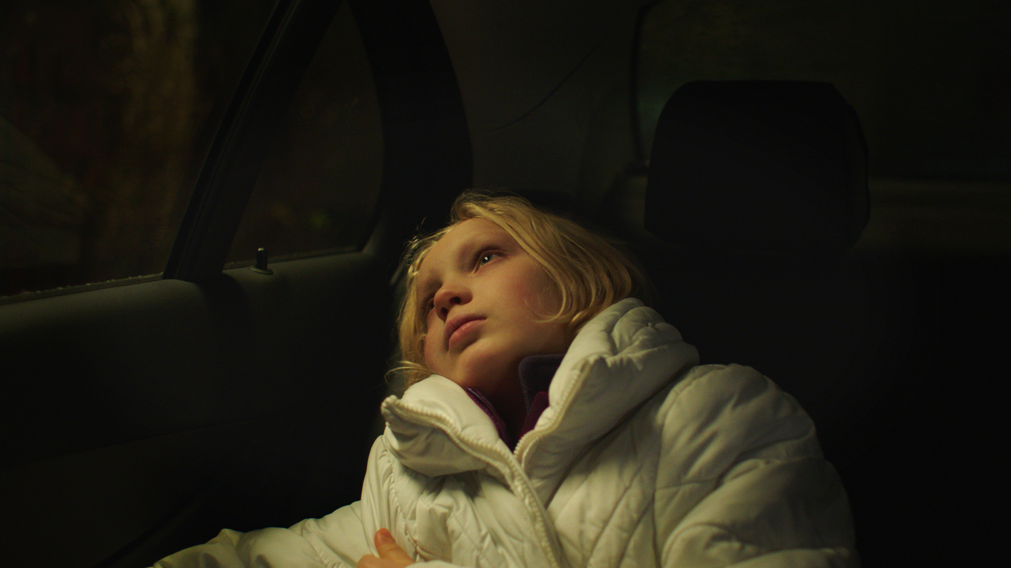 Kuvassa on nuori Helena Zengel, joka makaa puoliksi auton takapenkillä ja katsoo autosta ulos.  Hänellä vaaleat puolipitkät hiukset ja päällään valkoinen toppatakki.  Tausta on tumma.   