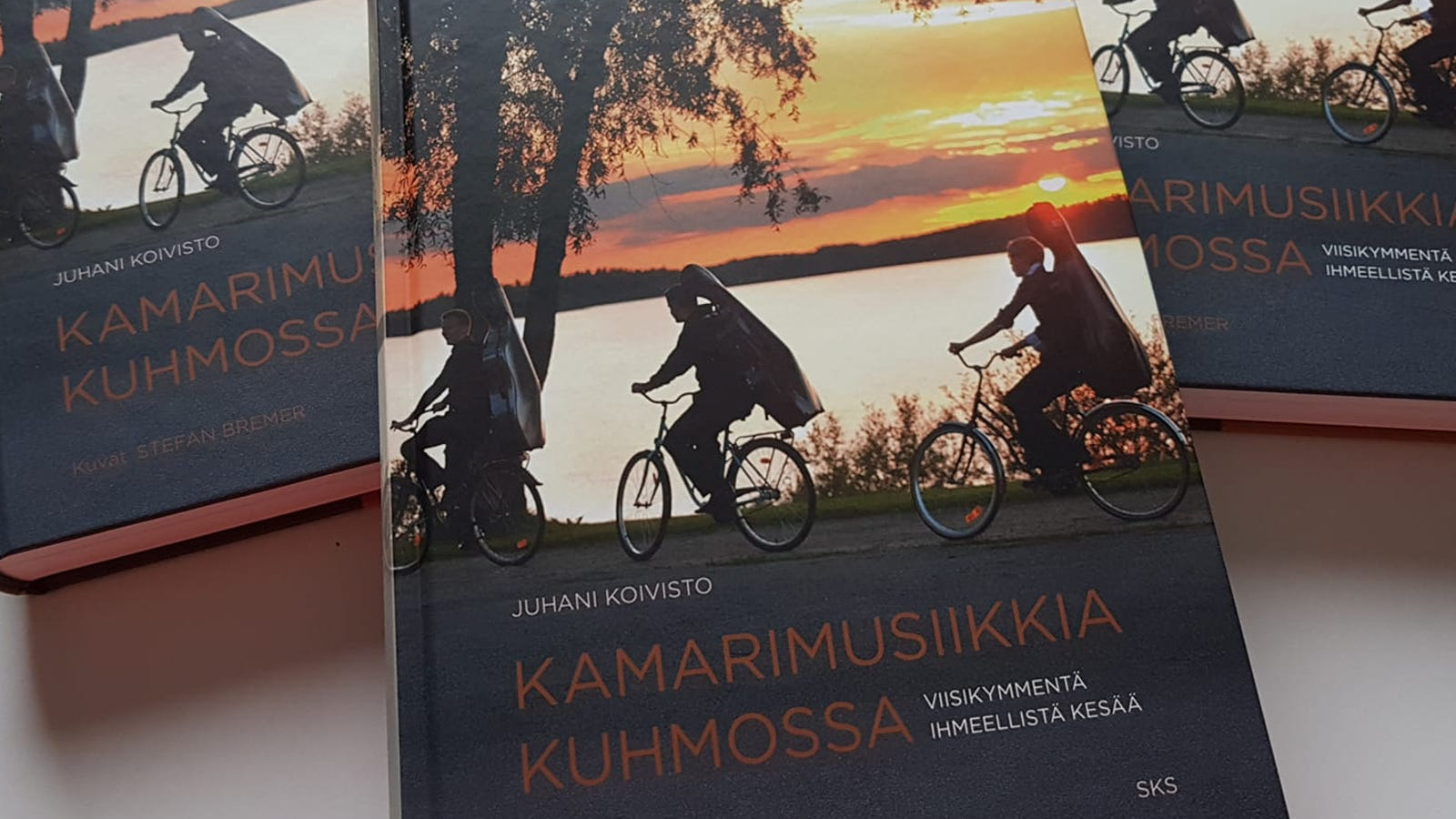 Kuvassa on kolme historiikkia, joiden kannessa on kuva kolme pyöräilevää soittajaan soittimet selässään.  Taustalla on järvi ja ilta-aurinkoinen taivas.  Kannessa lukee Kamarimusiikkia Kuhmossa, Viisikymmentä ihmeellistä kesää ja kirjoittaja Juhani Koivisto. Kuvat: Stefan Bremer. 