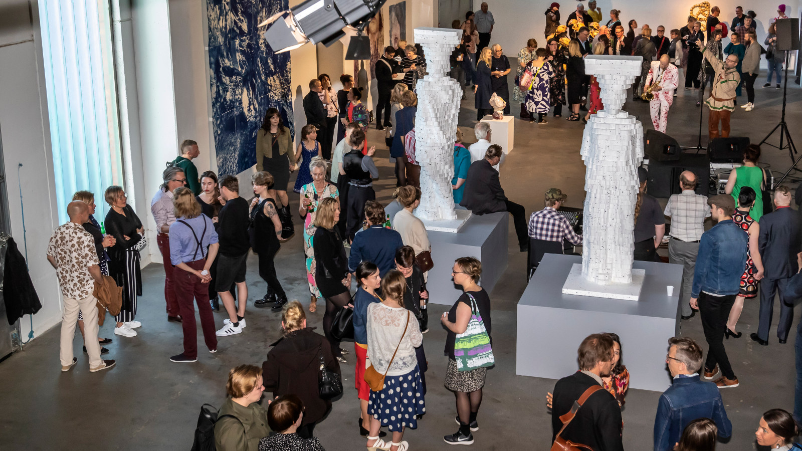 Kuvassa on ylhäältä päin kuvattu yleisöä Pekilossa Mäntässä.  Yleisöä on koko salin alalla pienissä ryhmissä keskustelemassa.  Salissa on valkoisia isoja patsaita.