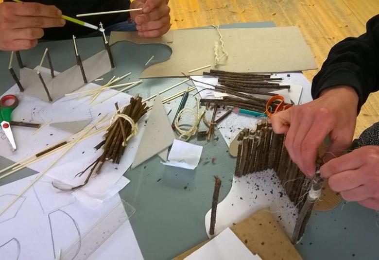 Kuvassa askarrellaan paperin ja lyhyiden oksanpätkien kanssa, jotka on leikattu noin 15-20 cm pitkiksi.  Vasemmassa ylänurkassa näkyy kädet ja menossa on oksanpätkiin valkoisten tikkujen liimaaminen ja tekeillä esteradalta näyttävä käytävä.  Oikeassa alareunassa näkyy toiset kädet, jotka kokoavat oksanpätkistä aitaa.  Pöydällä on pienet, vihreät sakset ja A4 papereita, joihin on piirretty luonnoksia ja oksanpätkiä.  