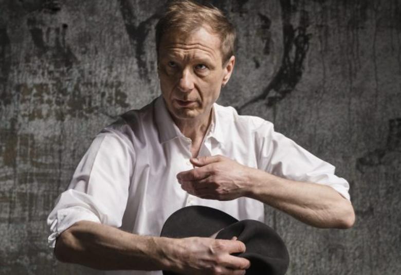 Kuvassa on Ismo Alanko valkoisessa paidassa hihat käärittyinä kyynerpäihin.  Oikeassa kädessä on musta hattu ja vasen käsi on oikean käden yläpuolella.  Tausta on ruskean musta ja kuvan sävy ruskean mustan valkoinen.