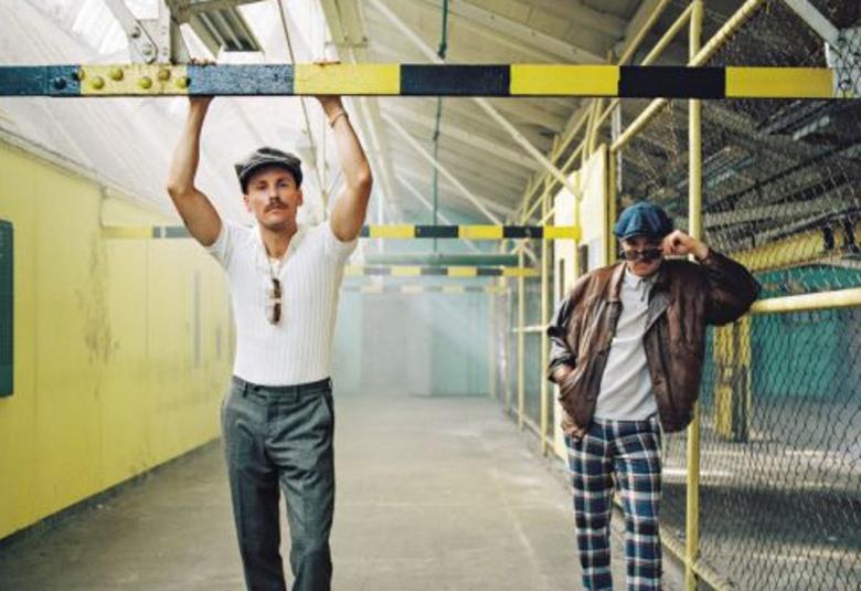 Kuvassa JVG:n jäsenet Jare ja VilleGalle ovat varastotilassa.  Etualalla on Jare valkeassa t-paidassa ja harmaissa, suorissa housuissa. Hänellä on kädet ylhäällä ja pitää kiinni kelta-mustasta palkista.  VilleGalle nojaa varastohäkin rautaverkkoon ja hänellä on yllään sini-mustaruudulliset housut, harmaa paita ja ruskea nahkatakki.  He seisovat varastokäytävässä, josta roikkuu kelta-mustia palkkeja ja varastohäkit ovat tyhjiä.