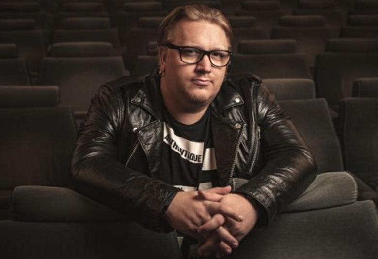 Kuva on Arttu Wiskarista, joka on istumassa tyhjässä katsomossa. Siellä on tummat penkit ja Wiskari nojaa edessä oleviin istuimiin kädet ristittyinä. Wiskarilla on musta nahkatakki päällään ja musta t-paita, jossa on valkoinen raita. Wiskarilla on silmälasit ja taaksepäin vedetyt ruskeat hiukset.