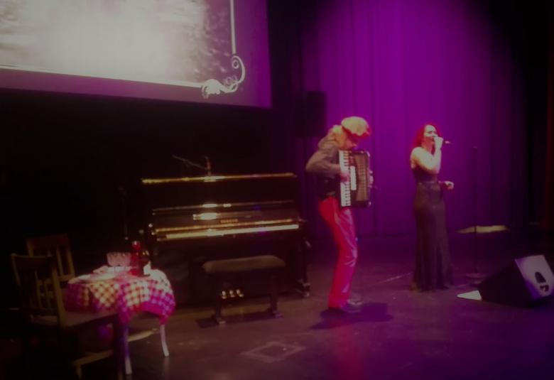 Kuvassa violetteisävyisessä näyttämötilassa oikella laulaa Minna Vainionpää, jolla on punainen pitkä tukka ja päällään ruskea, pitkä puku.  Hänen vasemmalla puolellaan on hanuria soittava Esko Grundström, jolla punainen nauha pään ympäri kietaistuna vaaleissa pitkissä hiuksissaan.  Hanuristin takana on piano pianon edessa vasemmalla on matala, valkoinen pöytä, jonka päällä valko-punaruutuinen liina. Vasemmalla ylhäällä on scriini, jolla on vaalea valoa. 