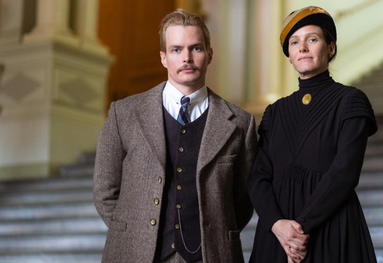 Kuvassa oikealla näyttelijä Johannen Holopainen ja vasemmalla Laura Birn.  Holopaisella on päällään harmaan ruskea puku, tumma napitettu liivi ja valkoinen paita.  Birnilla on päällään musta puku, jossa on ruskea medaljonki ja päässä keskiruskea baskeri.  Takana on harmaat portaat ja vasemmalla on klassinen pylväs ja portaikon takana vaalea kaari.