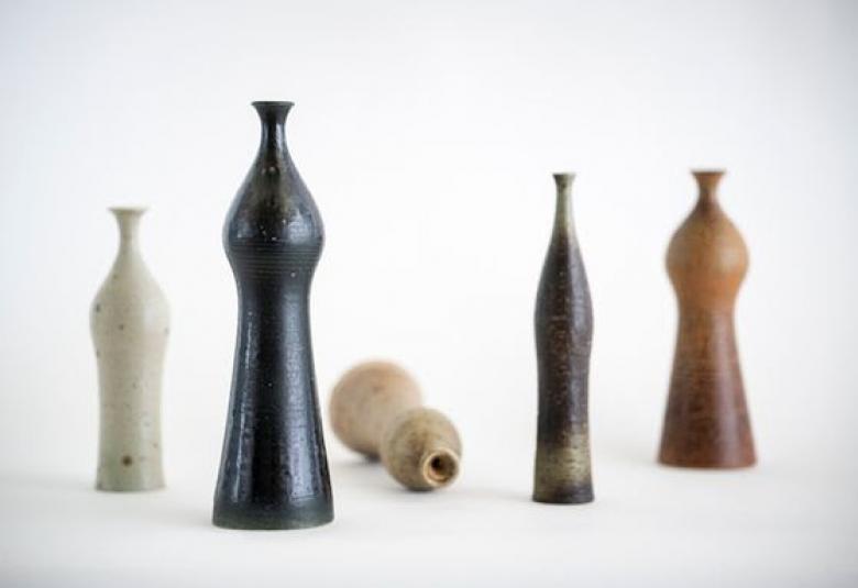 Kuvassa vaalealla taustalla eri kokoisia keramiikkapulloja. Vasemmalla vaalea, sitten isompi tumma, vaalea pitkällään maassa, tumma ja ruskea vieressä.