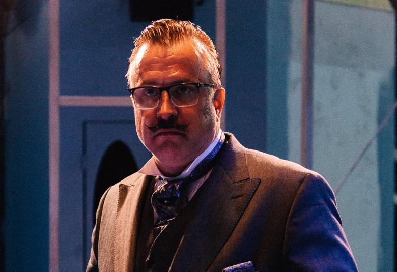 Kuva on Kristo Salmisesta, joka näyttelee Hercule Poirotia Suomenlinnassa. Salmisella on päällään tumma puvun takki ja valkoinen paita. Kuvassa näkyy hieman puvun kaulusta ja solmiota. Salmisella on päässään silmälasit ja vähän hiuksia päälaella sekä viikset.  Kasvot ovat hiukan varjossa.  Tausta on paikoin. tumman sininen 