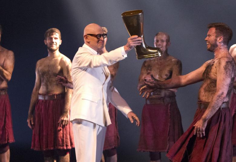 Kuvassa on Jorma Uotinen keskellä ja hän ojentaa saappaan seuraajalleen Riku Lehtipolulle.  Jorma Uotisella on päällään valkoinen puku.  Ympärillä on miestanssijoita, joilla on päällään ruskea hame ja paljas yläruumis, jossa on karvoitus.  Miehillä on paljaat jalat. 