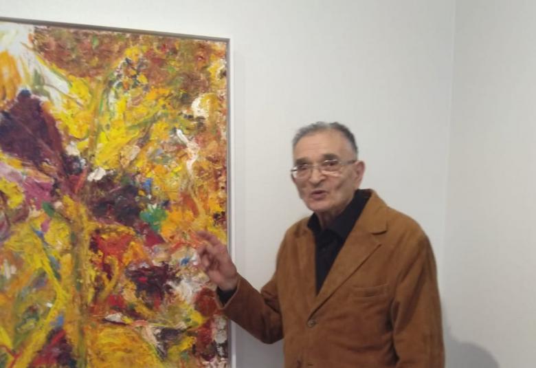 Kuvassa Rafael Wardi on oikealla ruskeassa takissa ja mustissa housuissa.  Hän osoittaa vasemmalla olevaa tauluaan, jonka nimi on Kukkia I.  Taulussa on oranssi pohja ja keskellä on isokukka, jossa on violetit terälehdet.