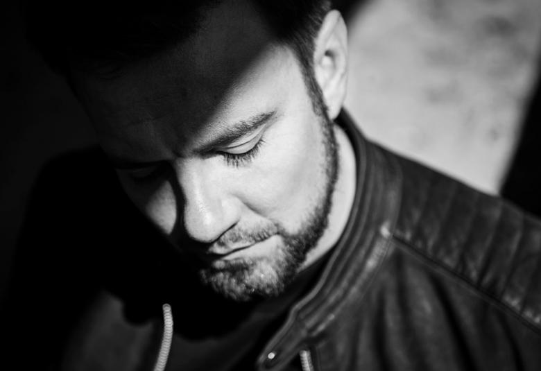 Kuva on Antti Ketosesta, joka katsoo alaspäin. Kuva on musta-valkoinen ja kasvokuva.  Kuvassa on valo tulee ylhäältä ja valaisee suurimman osan kasvoista.  Valojuova on myös taustalla.  Kuvassa näkyy hiukan mustan nahkatakin pystykaulusta.